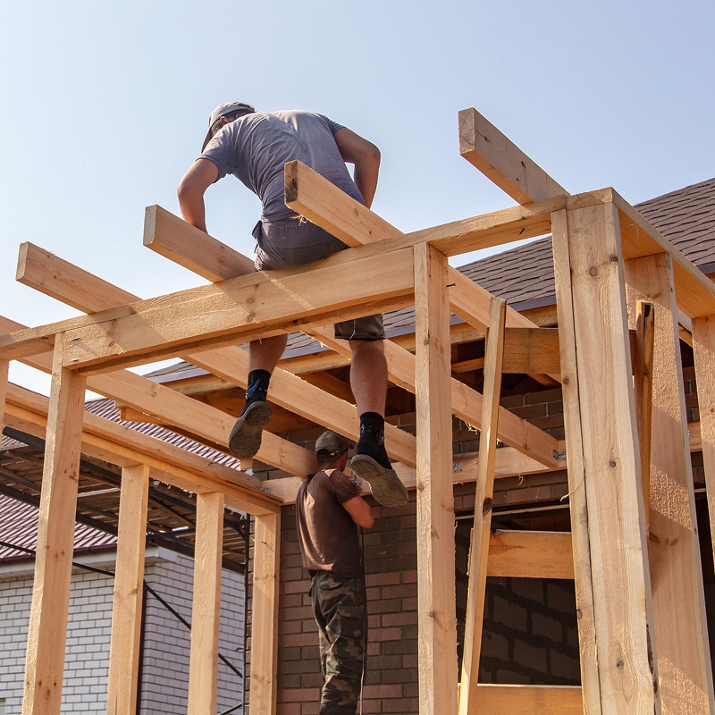Deux charpentiers construisent une ossature en bois pour une maison neuve sous un ciel clair. L'un est sur les poutres, l'autre en bas.