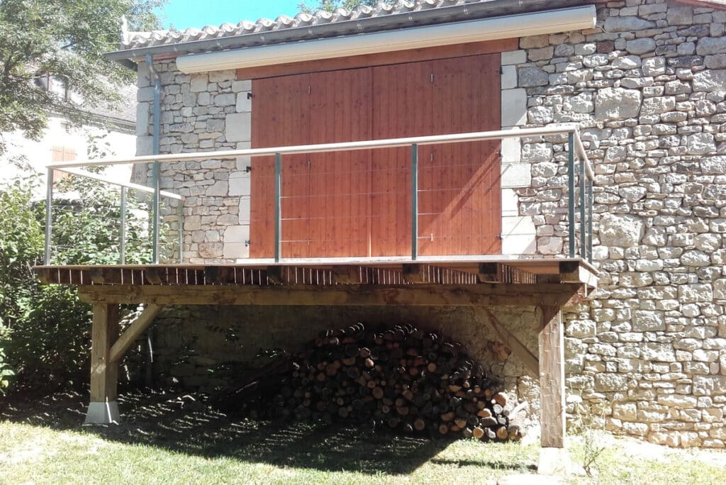 Balcon en bois et rambarde métallique sur façade de maison en pierre traditionnelle. Grande porte en bois, bûcher sous la terrasse.