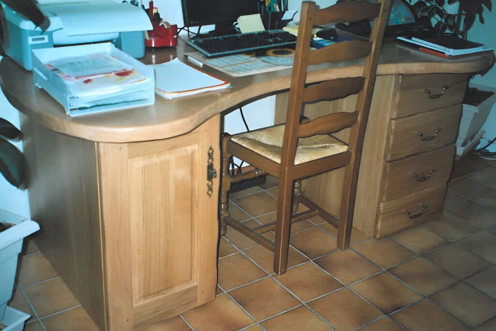 Bureau en bois clair avec un ordinateur, clavier, imprimante et plateau à documents. Une chaise en bois paillé est devant le bureau sur un sol carrelé.