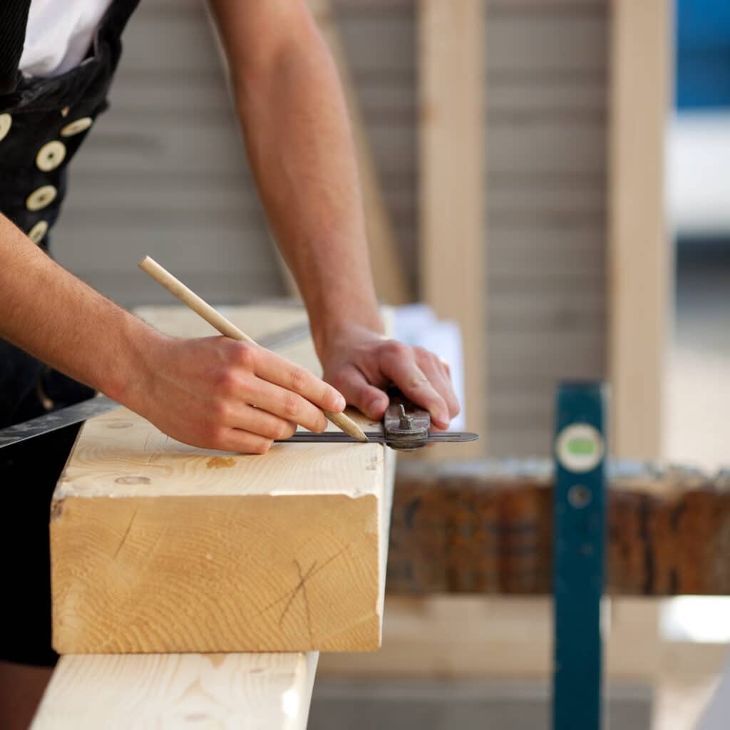 Mains d'artisan mesurant et marquant une poutre en bois clair avec un crayon et une jauge. Vêtement de charpentier traditionnel visible.