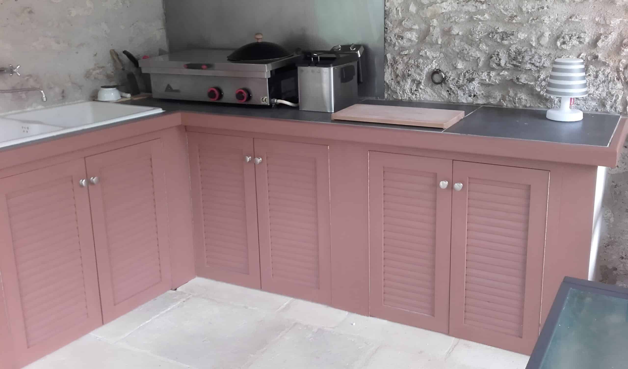Cuisine extérieure rustique avec meubles roses à persiennes, plan de travail gris, évier blanc, plancha et mur en pierre.