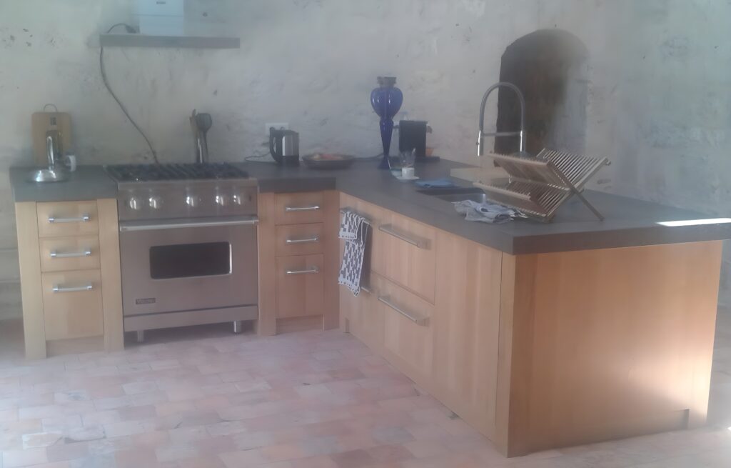 Cuisine rustique moderne en L avec murs en pierre, comptoirs gris, meubles en bois clair, cuisinière inox Viking et carrelage.