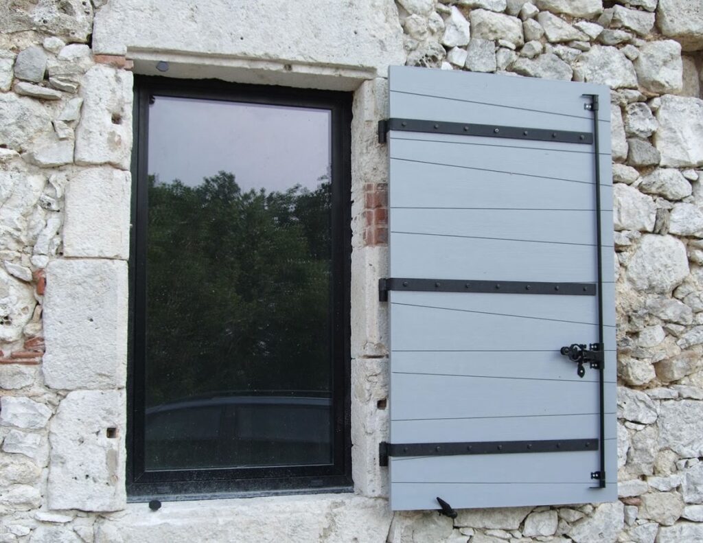 Fenêtre moderne noire dans mur de pierres brutes, un volet gris clair en bois et métal. Reflet d'arbres et de voiture sur la vitre.