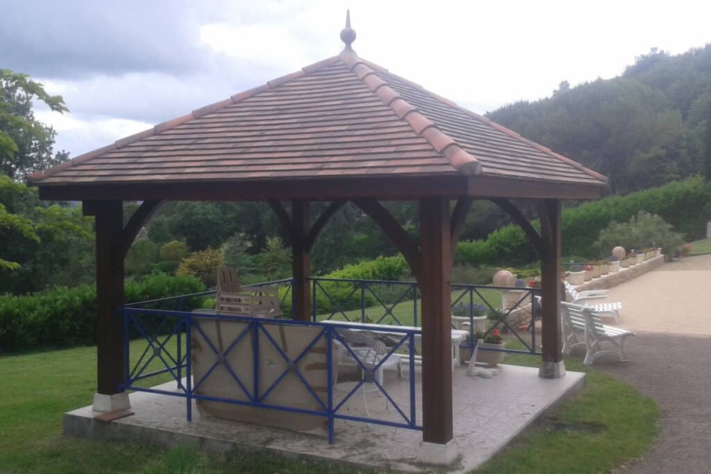 Grand gazebo en bois avec toit en tuiles ocres sur une terrasse pavée, entouré d'un jardin luxuriant et de mobilier de patio.