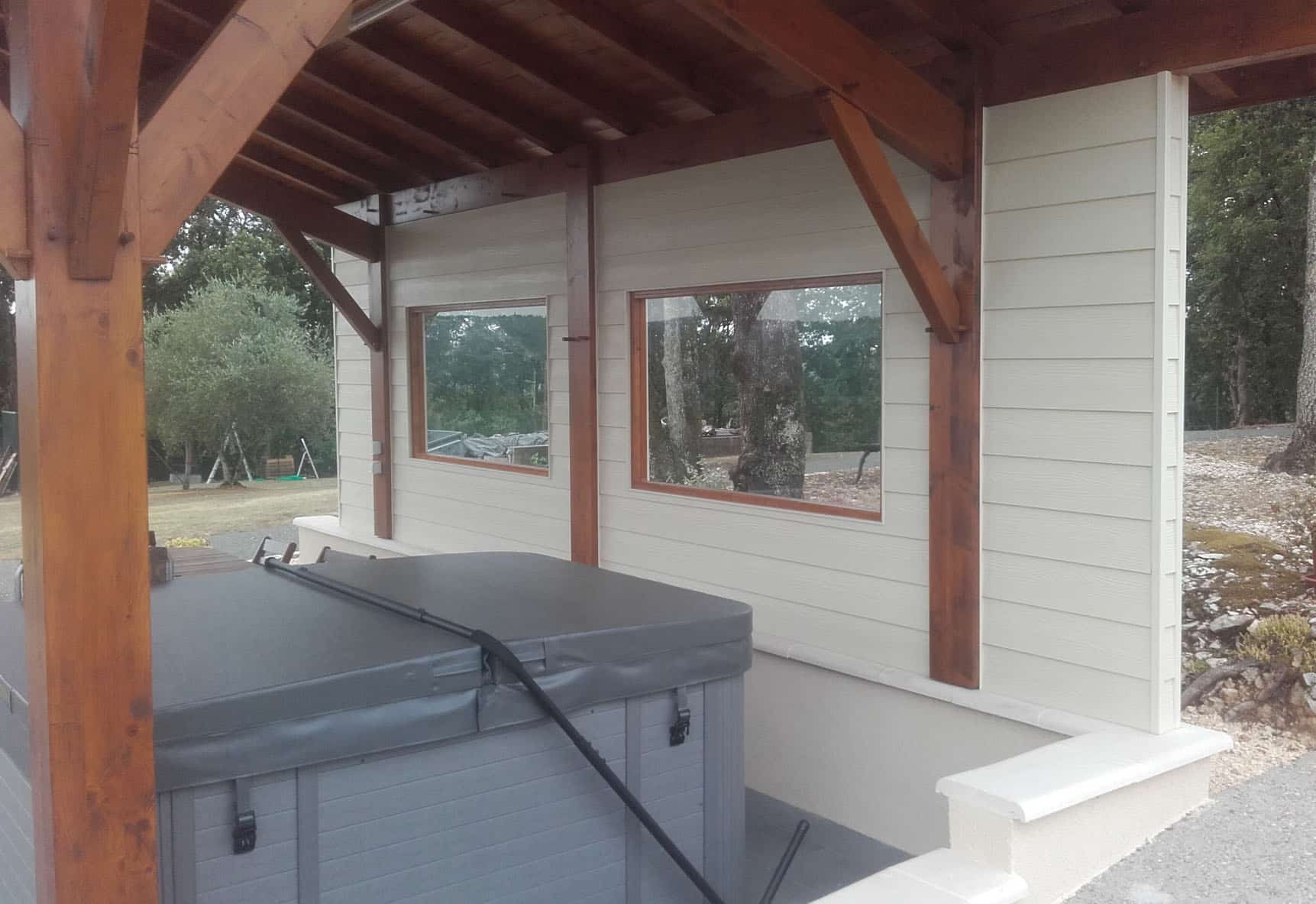 Patio en bois avec bardage clair et deux fenêtres, abritant un jacuzzi gris. Vue sur le jardin arboré.