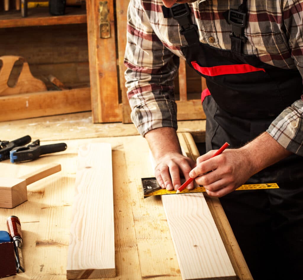 Menuisier marquant une planche de bois avec un crayon rouge et une règle métallique. Outils et établi en bois en arrière-plan, artisanat.