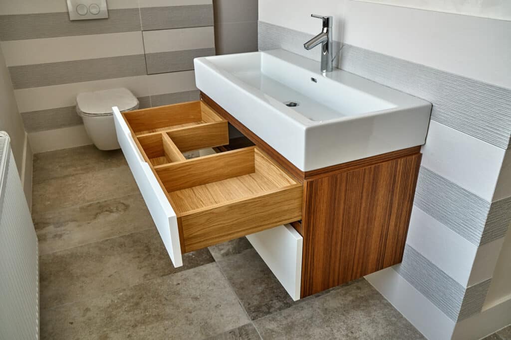 Salle de bain moderne: meuble vasque bois flottant, tiroir blanc ouvert avec rangements. Évier rectangulaire, carrelage mural rayé, sol clair.
