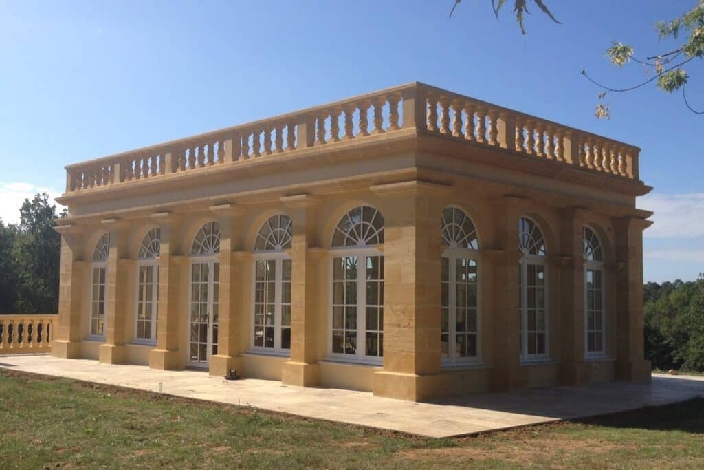 Pavillon classique en pierre blonde avec balustrade et grandes fenêtres cintrées blanches, entouré de verdure sous un ciel bleu.