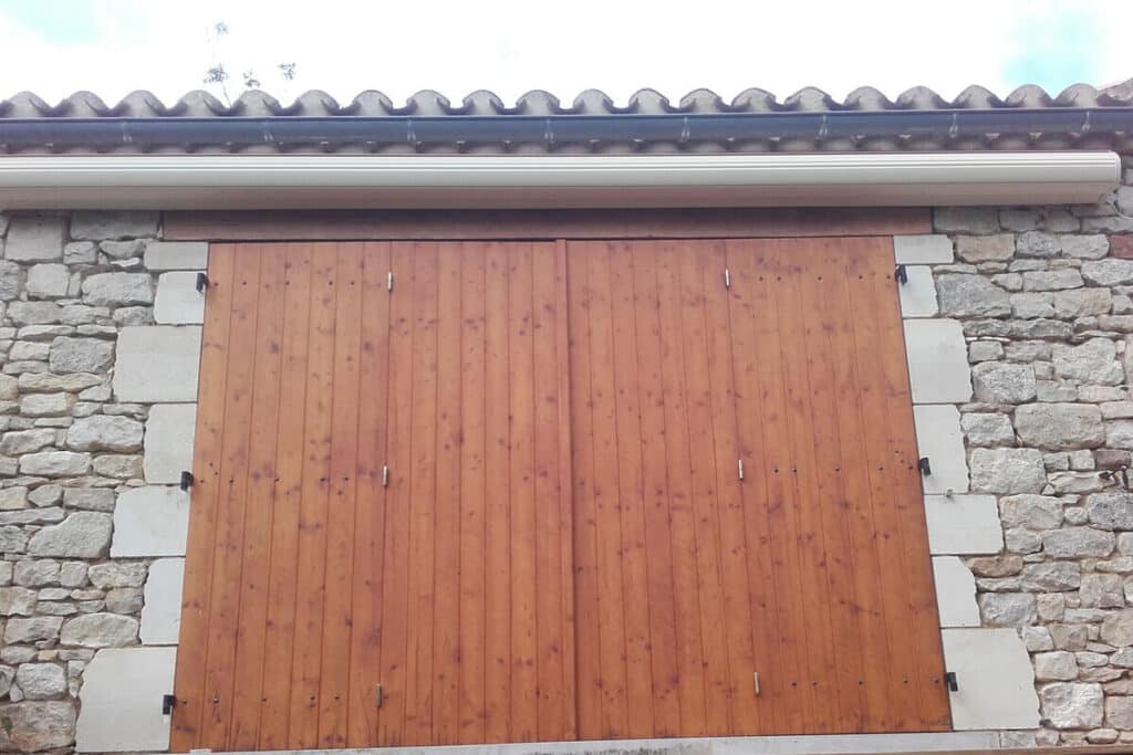 Grande porte ou volet en bois marron sur un mur en pierre naturelle, surmonté d'une gouttière claire et d'un toit en tuiles.