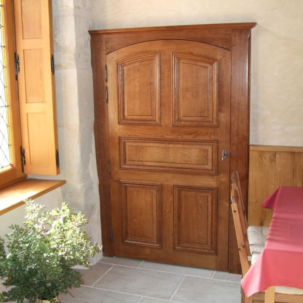 Grande porte en bois sculpté et fenêtres à volets dans une pièce rustique avec sol carrelé, plante de houx et table couverte.