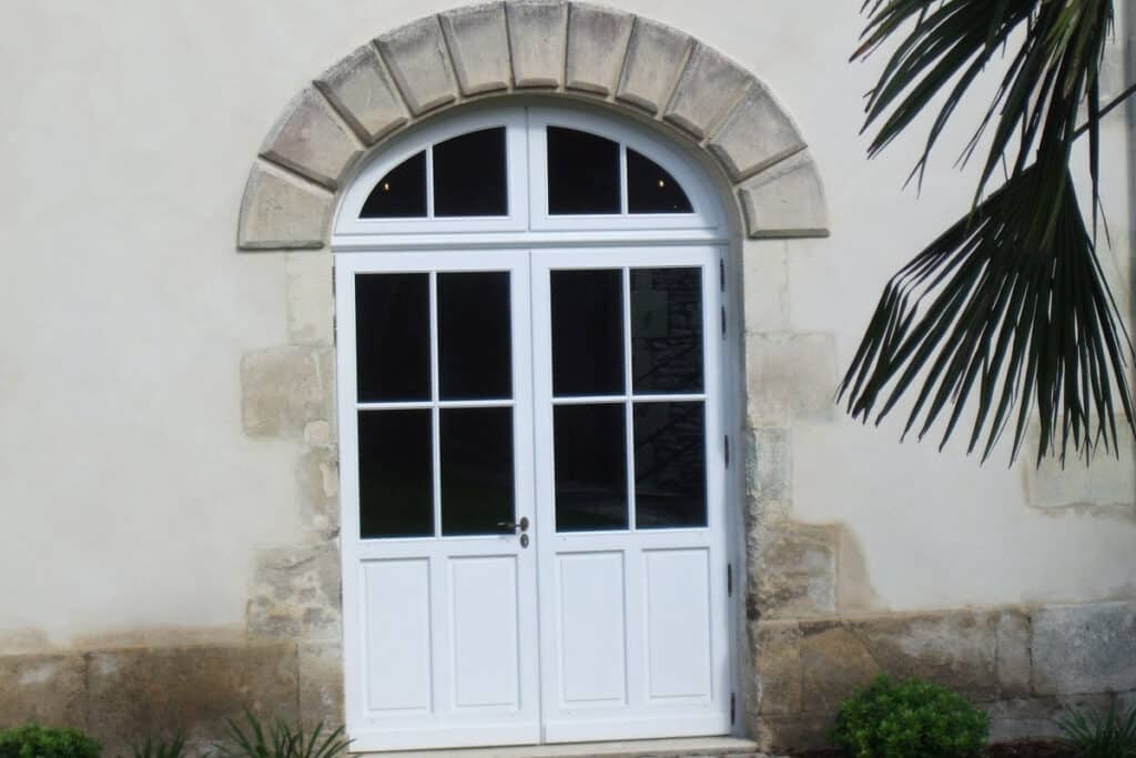 Double porte blanche vitrée sous un arc en pierre sculpté, sur façade claire. Feuille de palmier sur la droite.