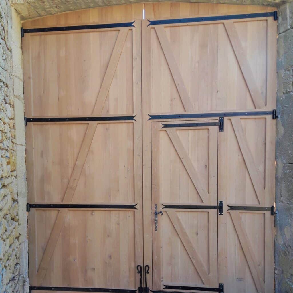 Grandes portes de grange en bois clair avec quincaillerie noire en fer forgé, incluant une porte piétonne, dans un mur de pierre rustique.