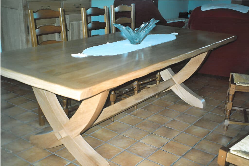 Grande table à manger en bois clair avec un piètement original en croix, ornée d'un centre de table en verre. La pièce est carrelée.
