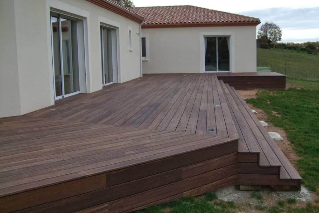 Vaste terrasse en bois foncé multi-niveaux avec escaliers, devant une maison beige et un jardin en pente. Accès par baies vitrées.