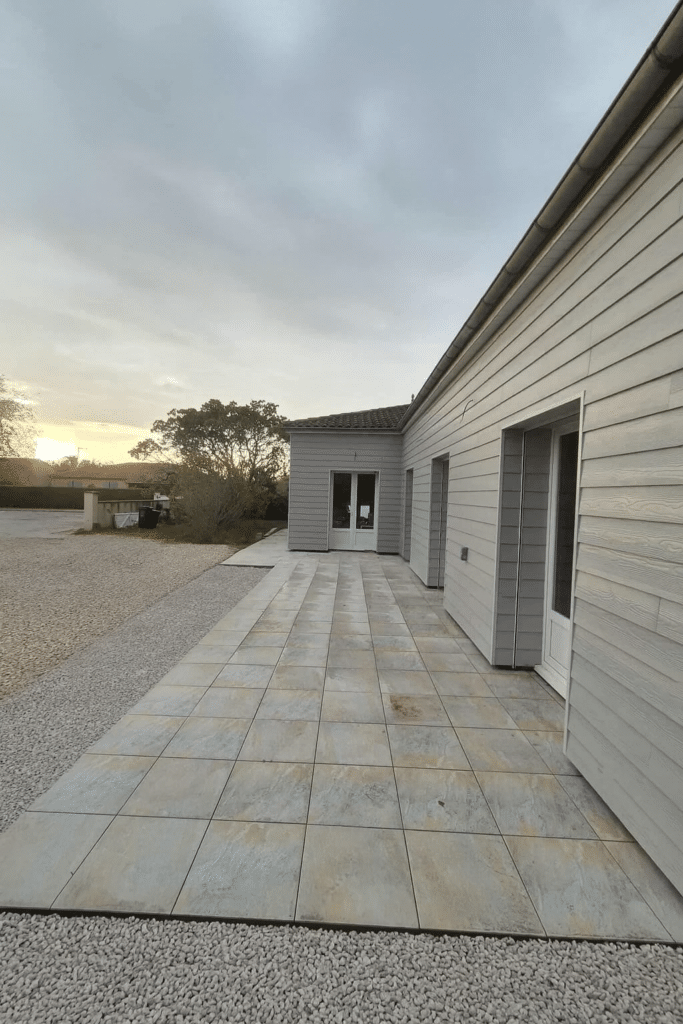 Terrasse carrelée en grès cérame devant une maison à bardage gris clair et gravier au coucher du soleil.